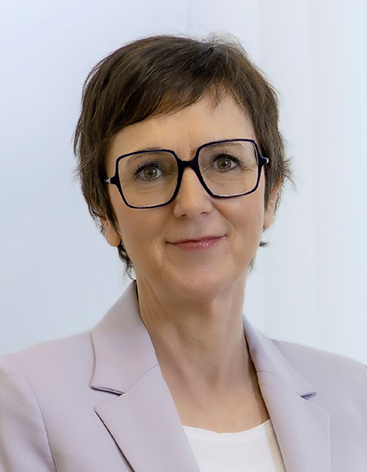 Porträt einer Frau mit kurzem Haar, Brille und hellem Blazer vor neutralem Hintergrund.